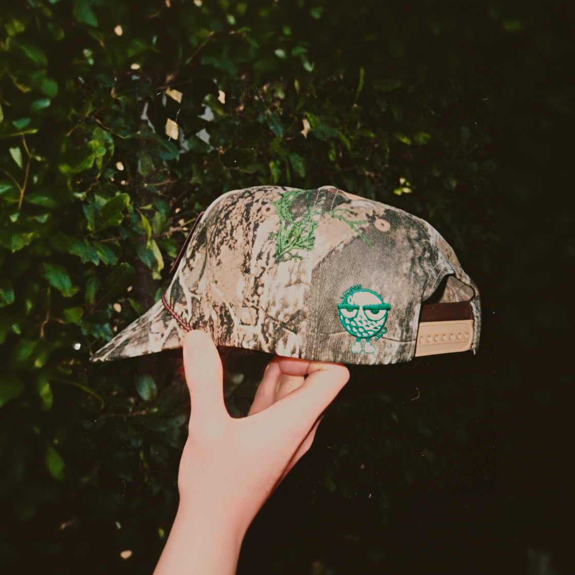A camouflage cap held by a hand against a dark green leafy background. The cap features a green embroidered pine tree design on the front and a green embroidered fish-eye patch on the side.