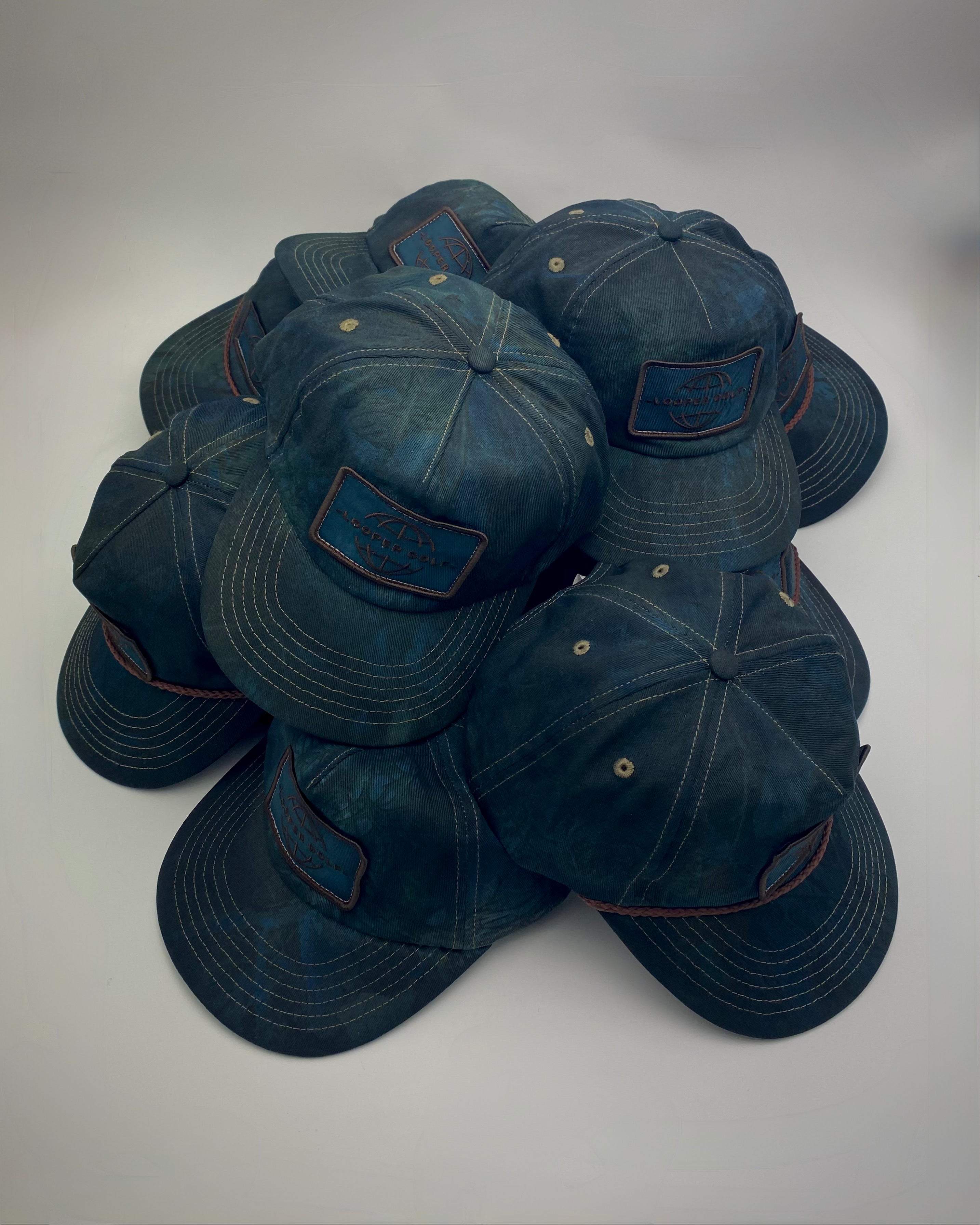 A pile of dark blue distressed caps with curved brims and a rectangular patch on the front, photographed on a light gray background.